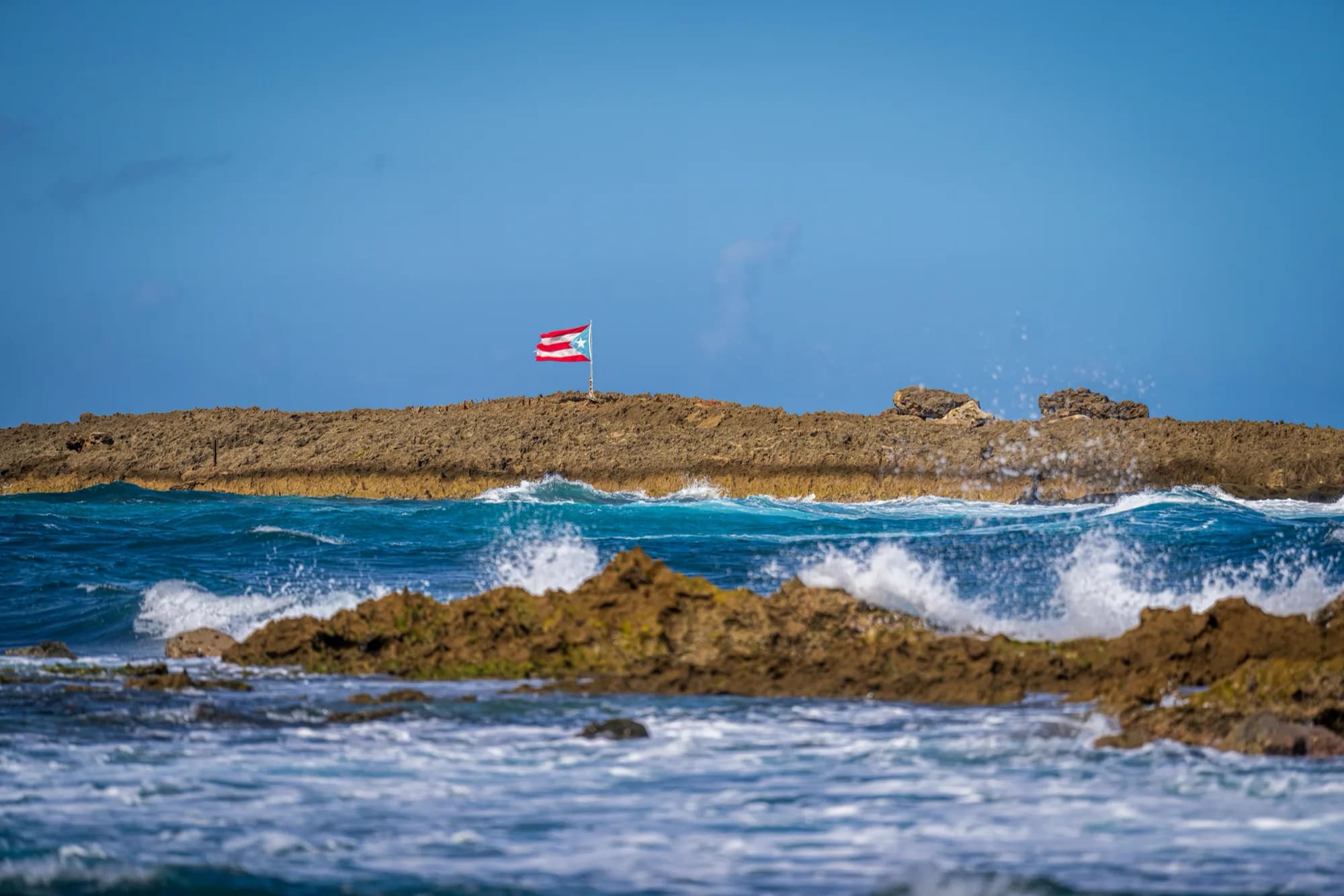 La bandera de Puerto Rico ondeando sobre una formación rocosa en la costa, con el oleaje rompiendo